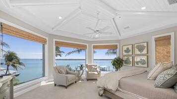 A bedroom with a large window overlooking the ocean, a bed, two chairs, and a ceiling fan.