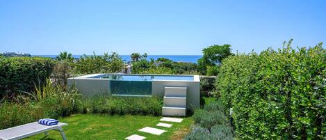 A modern outdoor pool area with a clear view of the sea, surrounded by lush greenery and a well-maintained lawn.