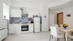 A modern kitchen with white cabinets, a blue and white tiled backsplash, a built-in oven, and a refrigerator. There is a dining table with chairs and a bowl of fruit on it. - TWO ROOMS SEA VIEW APARTMENT (FORIO)