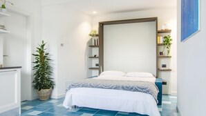 A bedroom with a bed, a bedside table, a nightstand, a wall-mounted shelf with decorative items, and a potted plant. - NEW DELUXE STUDIO WITH PRIVATE GARDEN AND TERRACE (Forio)