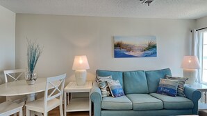 A living room with a teal sofa, white chairs, a small table, and a painting of a beach scene. - Grand Beach Resort #311 - Close to Beach with Gulf Views and Resort Amenities (Gulf Shores)