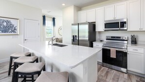 A modern kitchen with white cabinets, a stainless steel refrigerator, a microwave, and an oven. There are three bar stools with cushioned seats. - Margaritaville Fun in the Sun - Townhome (Kissimmee)