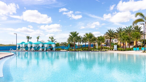 A swimming pool with lounge chairs and umbrellas, surrounded by palm trees and a building with a blue roof. - Margaritaville Fun in the Sun - Townhome (Kissimmee)