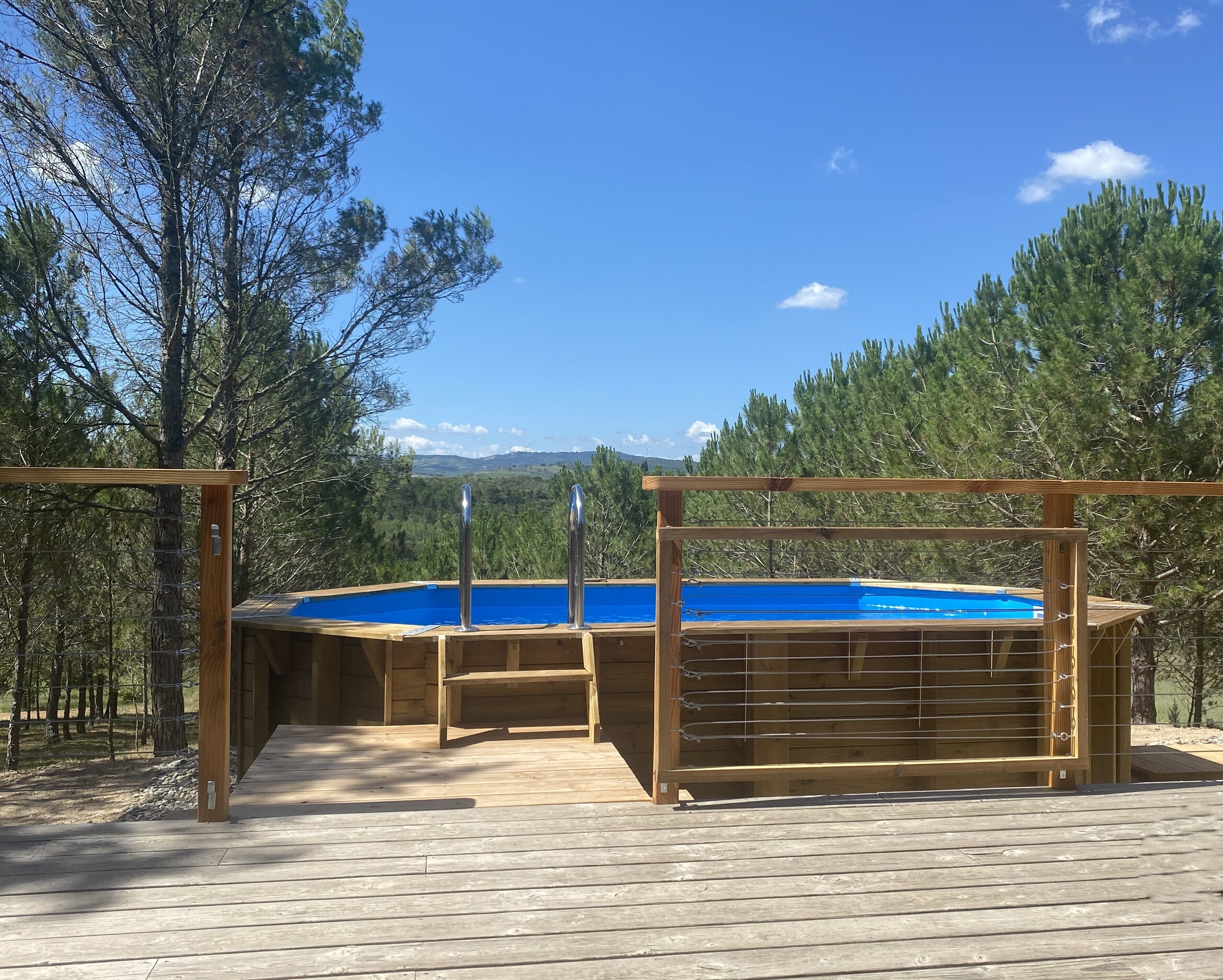 Una terraza de madera con una piscina azul, rodeada de árboles y un cielo despejado.