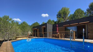Een modern buitenzwembad met een houten terras, een zwemtrap en een overdekt zitgedeelte met een rode parasol.