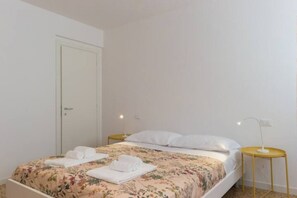 Un letto singolo con lenzuola a motivi floreali, un comodino con una lampada e una porta a sinistra.