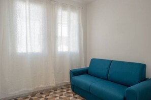 A room with a blue sofa, a window with sheer curtains, and a patterned floor.