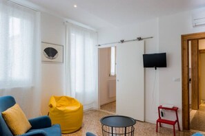 Une pièce avec une chaise bleue, un fauteuil poire jaune, une petite table ronde, un tabouret rouge et un téléviseur fixé au mur.