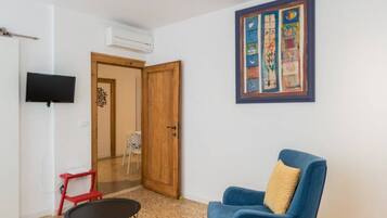 Una stanza con una porta in legno, una televisione fissata al muro, una poltrona blu con cuscino e un tavolino.
