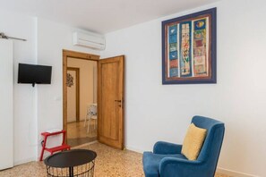Una habitación con puerta de madera, un televisor montado en la pared, una silla azul con cojín y una mesita.