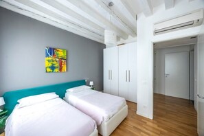 A room with two beds, a teal headboard, a white wardrobe, and a colorful abstract painting on the wall.