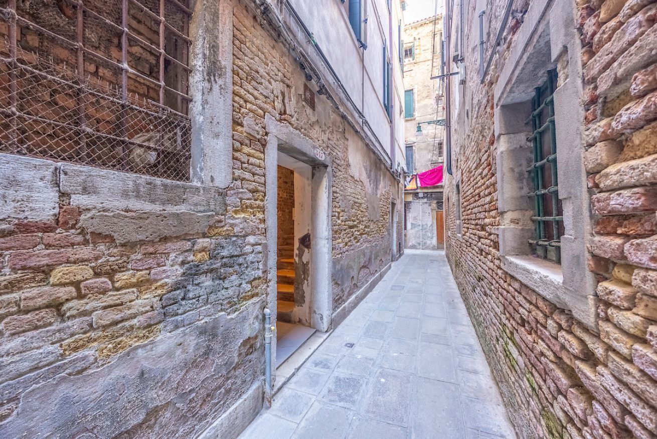 Un stretto vicolo con pareti in pietra e pavimento piastrellato, che conduce a un edificio con porta aperta.