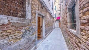 A narrow alleyway with stone walls and a tiled floor, leading to a building with an open door.