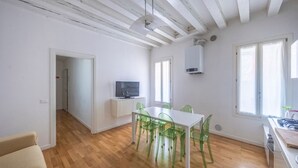 A room with a wooden floor, a white table with green chairs, a television on a stand, and a window with curtains.