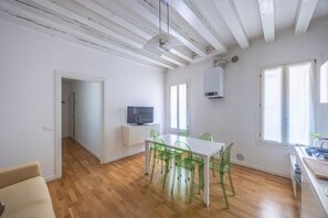 A room with a wooden floor, a white table with green chairs, a television on a stand, and a window with curtains. - Rialto al Manganer, apartment near Rialto Mercato (Venezia)