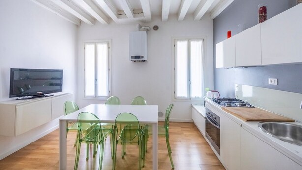 A modern kitchen with white cabinets, a gas stove, and a dining area with a white table and green chairs. - Rialto al Manganer, apartment near Rialto Mercato (Venezia)