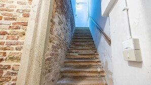 A narrow stairway with a brick wall on one side and a white wall on the other, leading to a door at the top.