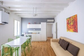 A modern kitchen with white cabinets, a dining table with green chairs, a beige sofa, and a colorful abstract painting on the wall.