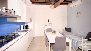 Una cocina compacta con armarios blancos, un protector antibacteriano azul y una estufa a gas. Hay una mesa de comedor con sillas y una planta pequeña encima. Se ve una cama con un edredón y almohadas a rayas al fondo.