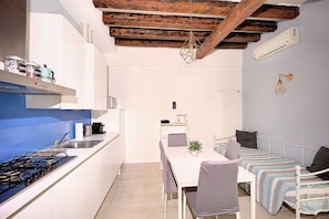 A compact kitchen with white cabinets, a blue backsplash, and a gas stove. There is a dining table with chairs and a small plant on it. A bed with a striped blanket and pillows is visible in the background.
