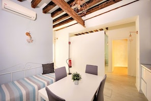 A dining area with a white table, grey chairs, and a striped bench. There is a potted plant on the table. The room has wooden beams on the ceiling and a fire extinguisher on the wall.