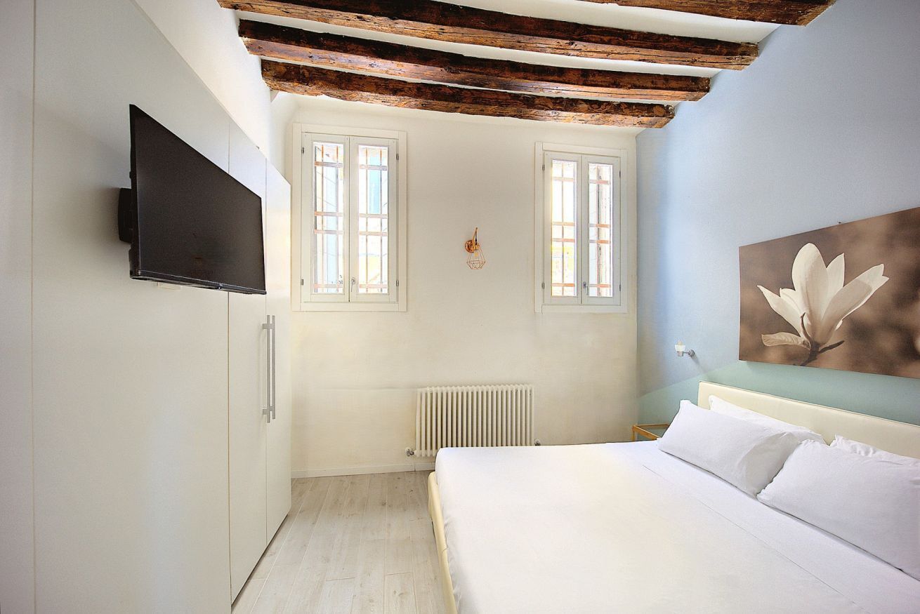 Ein Schlafzimmer mit Bett, Wandfernseher, zwei Fenstern, Heizkörper und einem Holzbalken an der Decke.