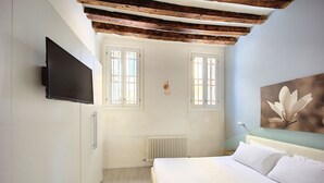 A bedroom with a bed, a TV mounted on the wall, two windows, a radiator, and a wooden ceiling beam.