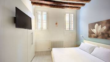 Ein Schlafzimmer mit Bett, Wandfernseher, zwei Fenstern, Heizkörper und einem Holzbalken an der Decke.