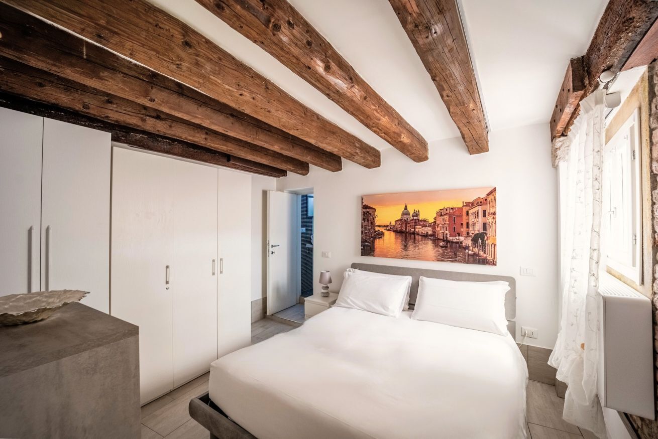 A bedroom with a bed, white wardrobe, and a painting of a cityscape.