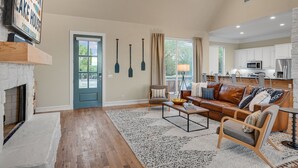A spacious living room with a fireplace, a large window, and a kitchen in the background.