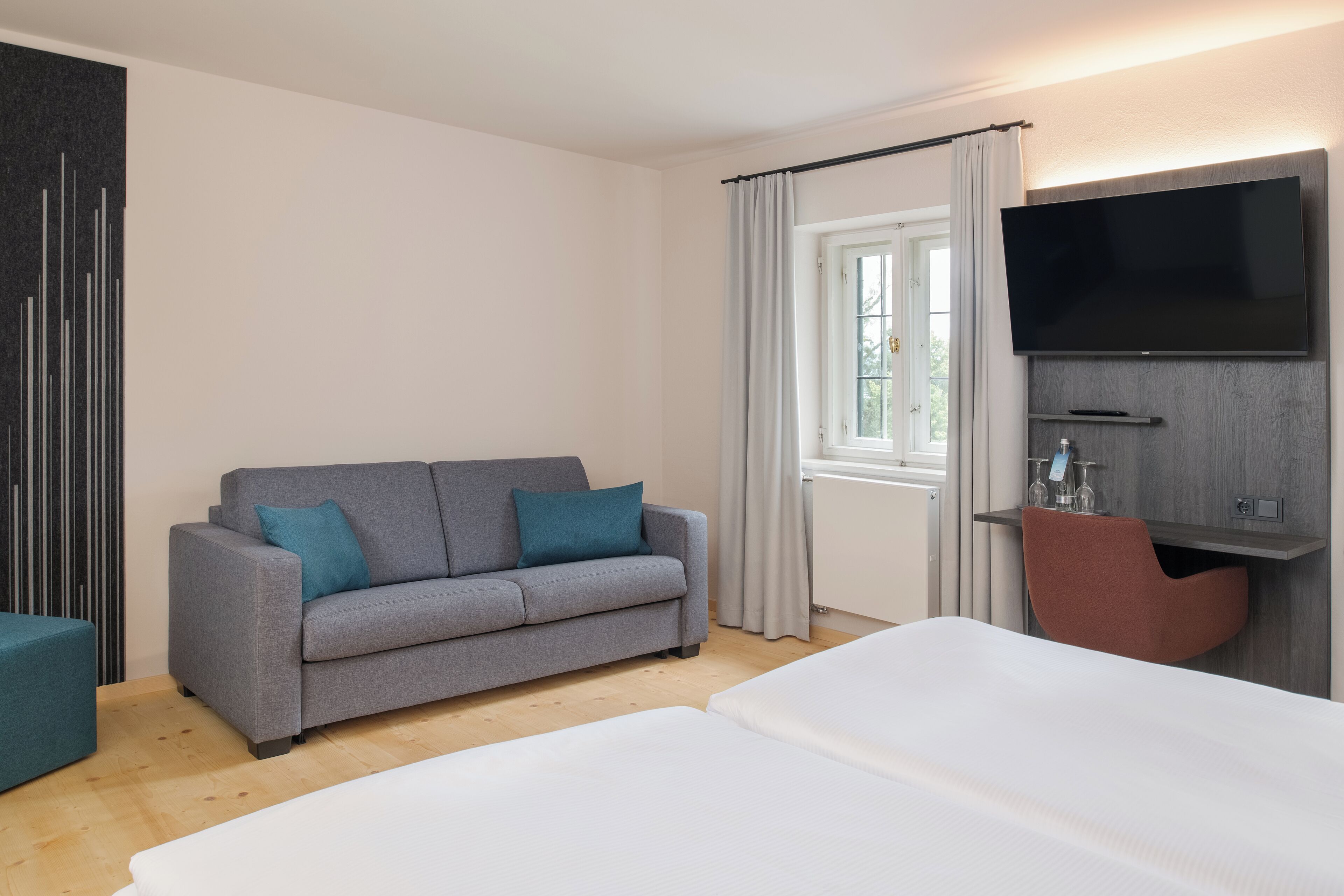 Una habitación de hotel moderna con sofá gris, cama blanca, televisor de pantalla plana en la pared y un pequeño escritorio con silla.