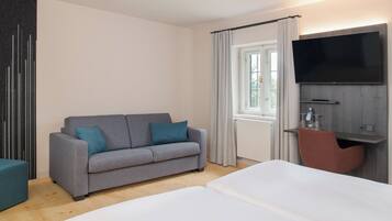 Una habitación de hotel moderna con sofá gris, cama blanca, televisor de pantalla plana en la pared y un pequeño escritorio con silla.