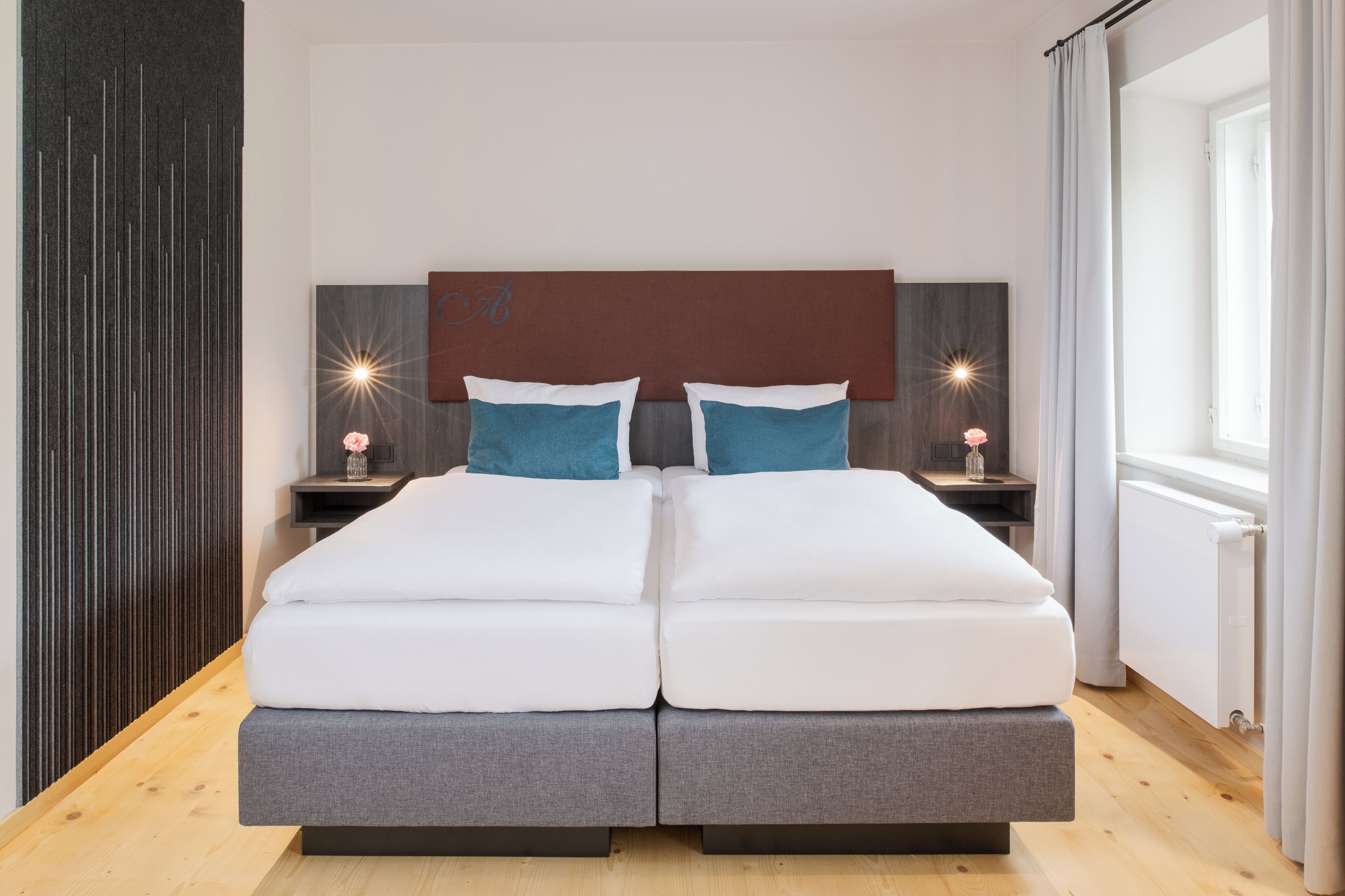 A double bed with a wooden headboard, two bedside tables with lamps, and a view of the outdoors through a window with curtains.