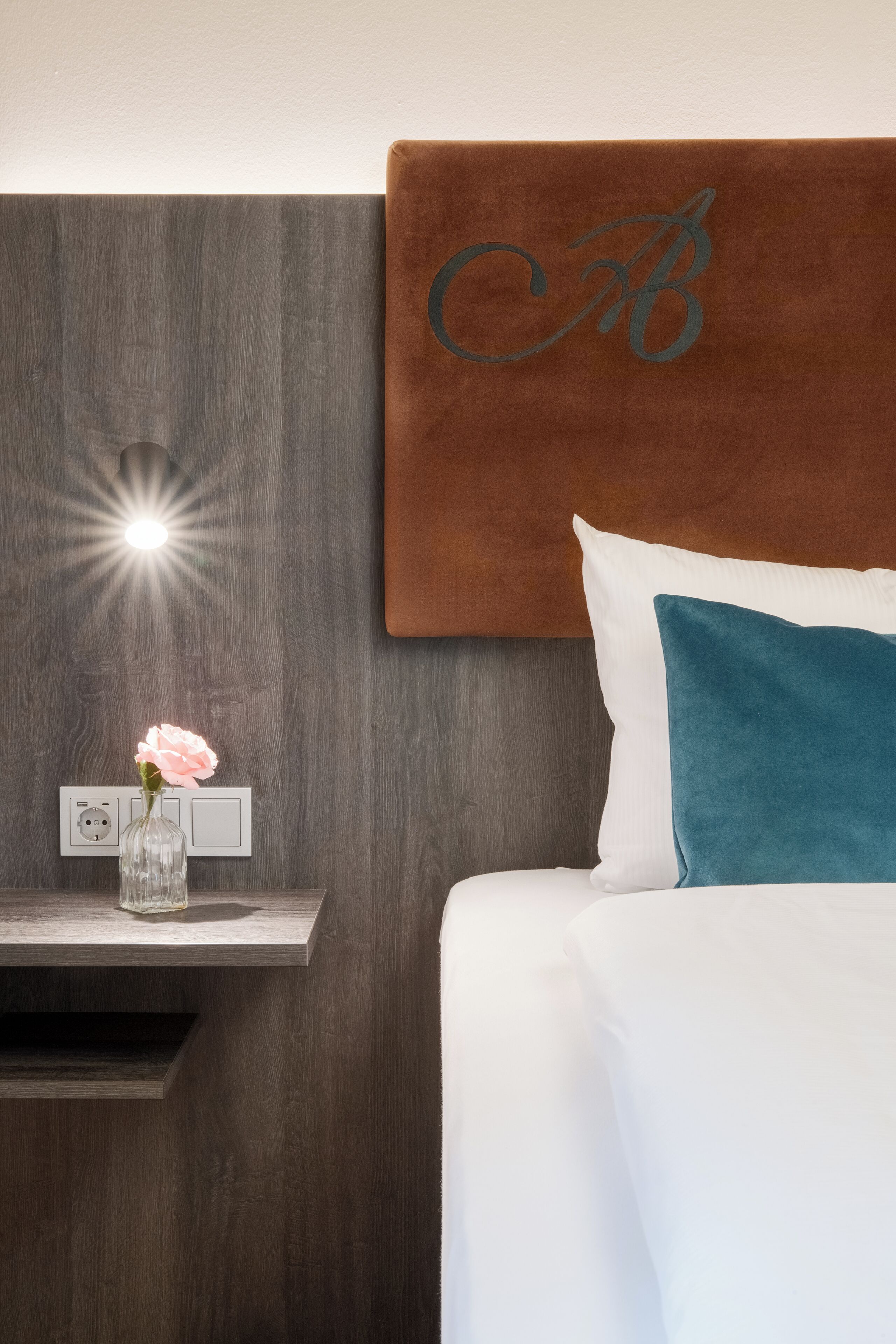 A neatly made bed with white and teal pillows, a wooden headboard with a monogram, a bedside table with a vase of flowers, and a wall socket.