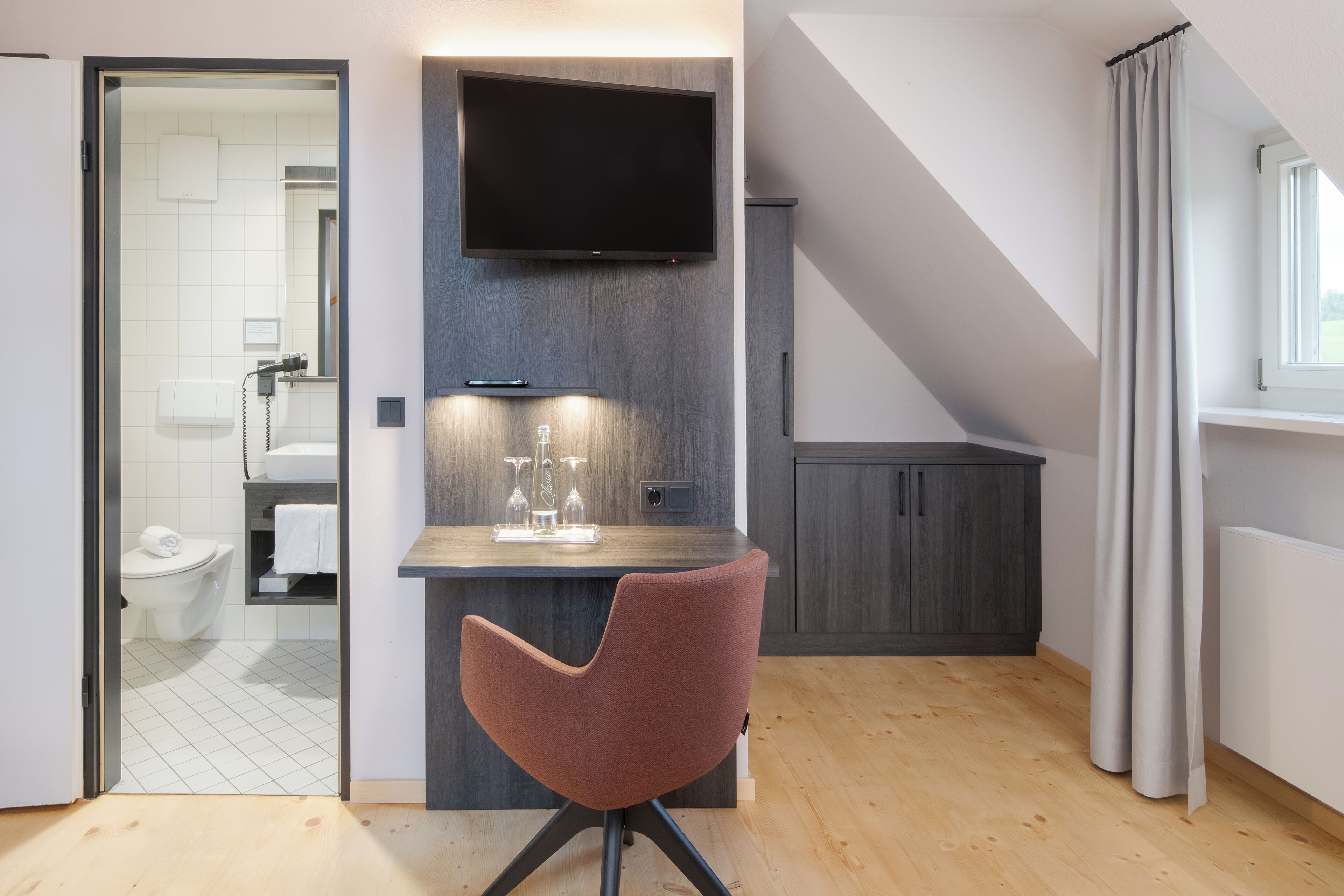 Ein modernes, kompaktes Wohnzimmer mit Holzboden, einem flachbildfernseher an der Wand, einer kleinen Küchenzeile mit Arbeitsplatte und Schränken, und einem durchgehenden Türspalt sichtbaren Badezimmer.
