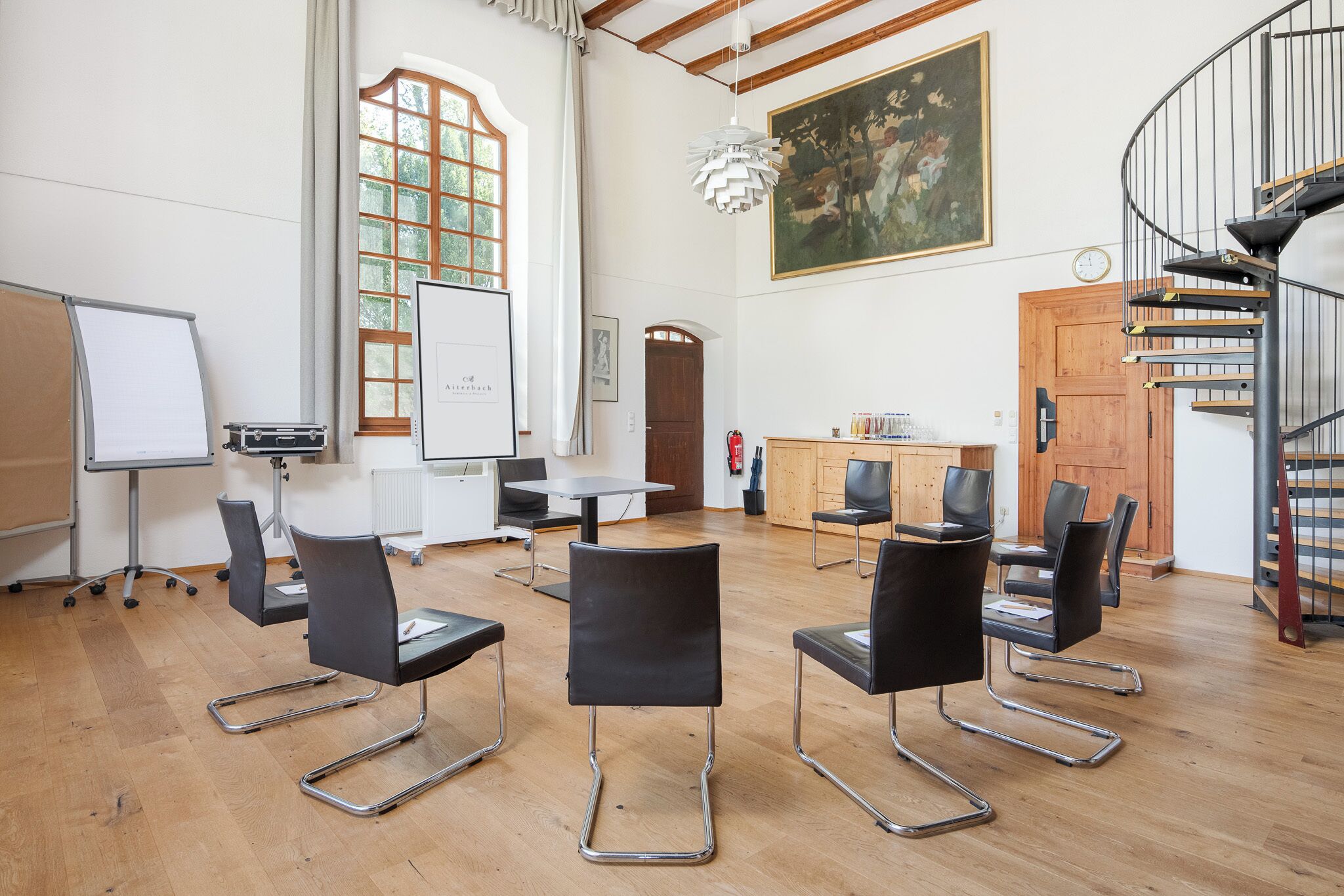 Ein geräumiger Raum mit einer Whiteboard-Tafel, schwarzen Stühlen, Holzboden, einem Gemälde und einer Wendeltreppe.