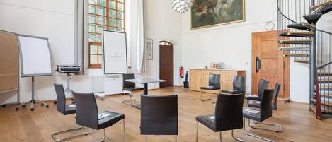 Ein geräumiger Raum mit einer Whiteboard-Tafel, schwarzen Stühlen, Holzboden, einem Gemälde und einer Wendeltreppe.