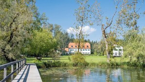 Un pont en bois enjambe une rivière paisible et mène à une grande maison avec un toit rouge.