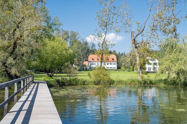 A wooden bridge over a calm river leading to a large house with a red roof.