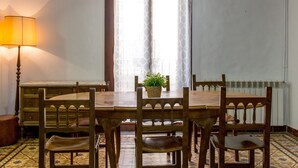 Un coin repas avec une table et des chaises en bois, une lampe, une fenêtre ornée d’un rideau à motifs et un radiateur.