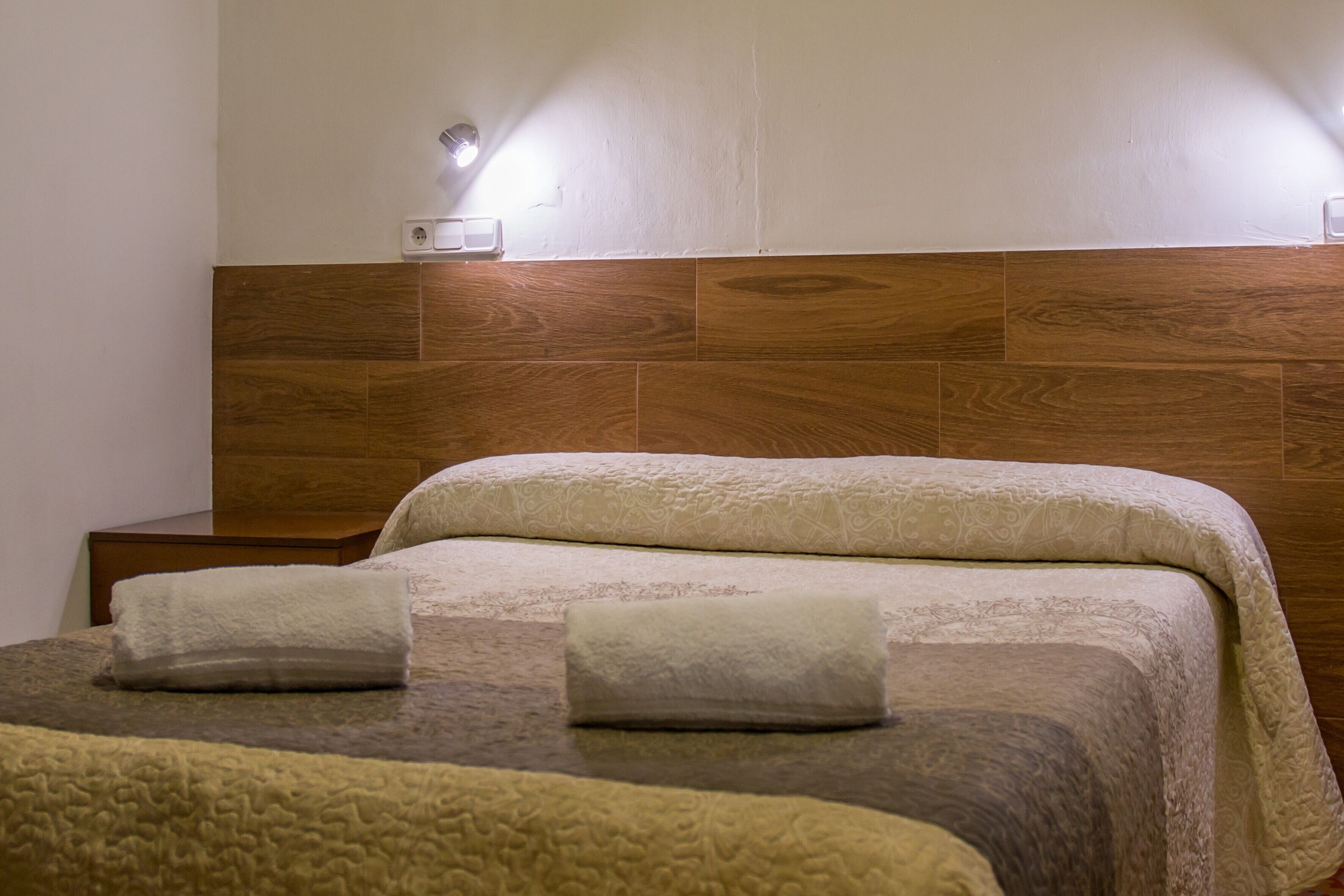 Une chambre d’hôtel avec une tête de lit en bois, deux lits avec une literie beige, une table de chevet et une lampe fixée au mur.