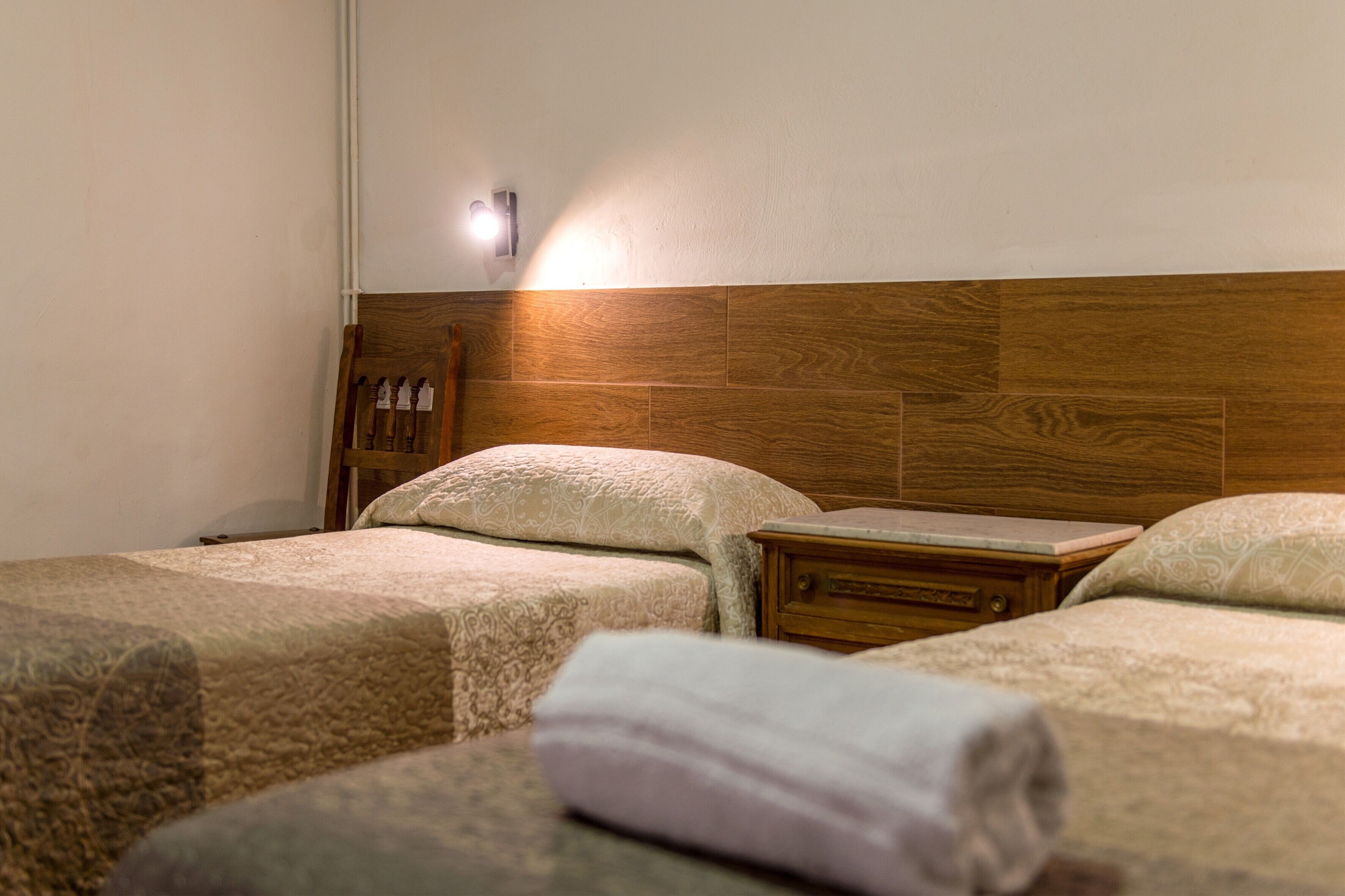 Habitación de hotel con dos camas, cabecero de madera, mesita de noche y lámpara de pared.