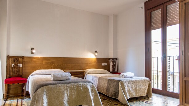 Une chambre d’hôtel avec deux lits, une chaise rouge, une tête de lit en bois, un sol carrelé et un balcon avec une balustrade.