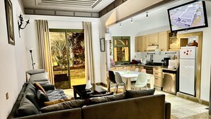 A modern living room with a sofa, dining table, and chairs. There is a kitchen area with cabinets and appliances. A television is mounted on the wall. - Au Coeur De La Palmeraie De Marrakech  (Marrakech)