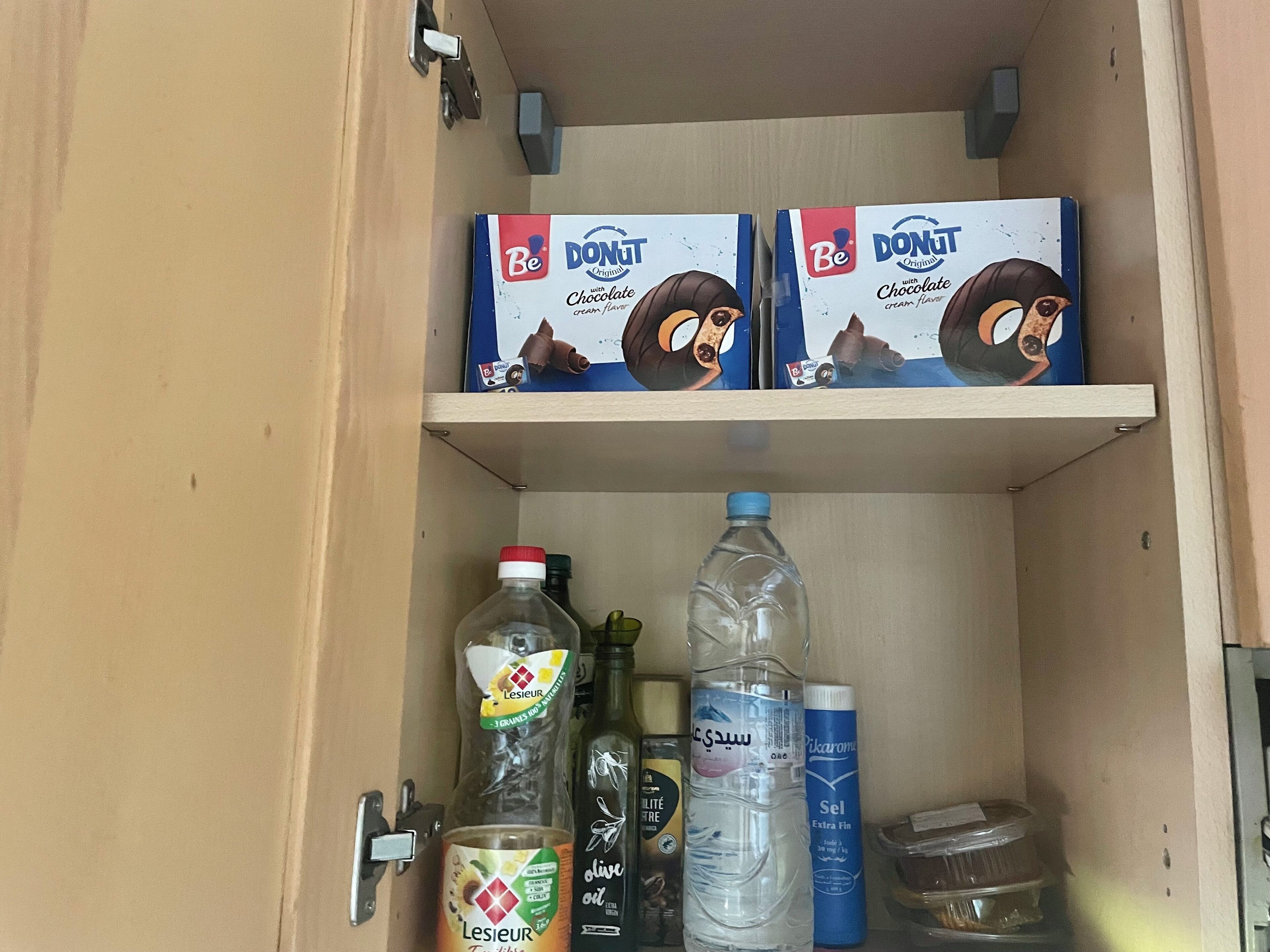 A cabinet with two boxes of Be Donut Original Chocolate Cream flavor, a bottle of olive oil, a bottle of water, a bottle of Lesieur Tinctures, and a container of Sel Extra Fin.