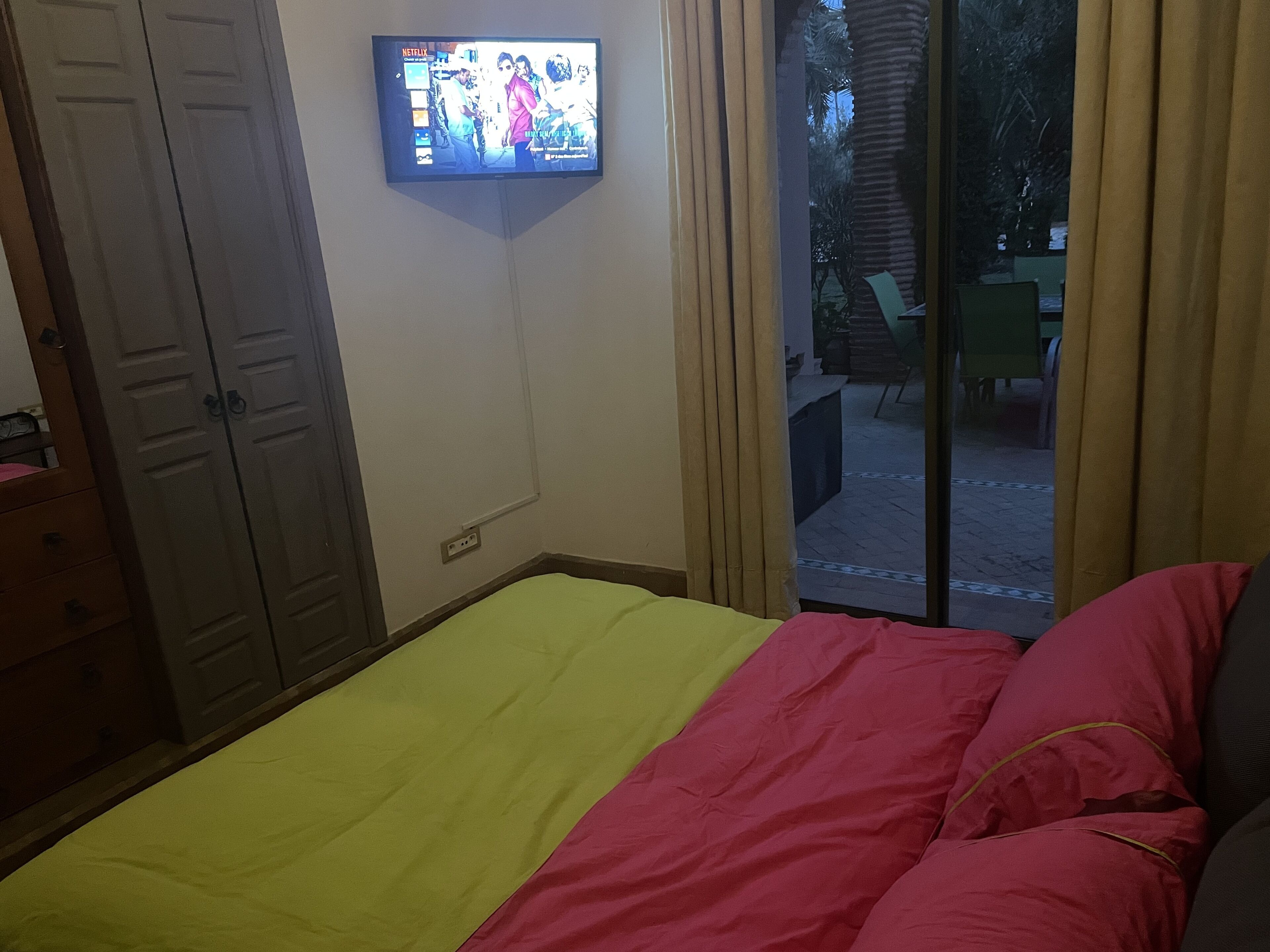 A bedroom with a bed, a TV mounted on the wall, a wardrobe, and a sliding glass door leading to an outdoor area.