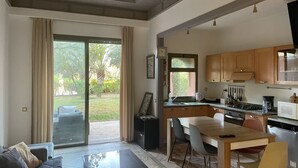 A spacious living area with a sliding glass door leading to a garden, a dining table with chairs, and a kitchen with wooden cabinets and a stove. - Au Coeur De La Palmeraie De Marrakech  (Marrakech)