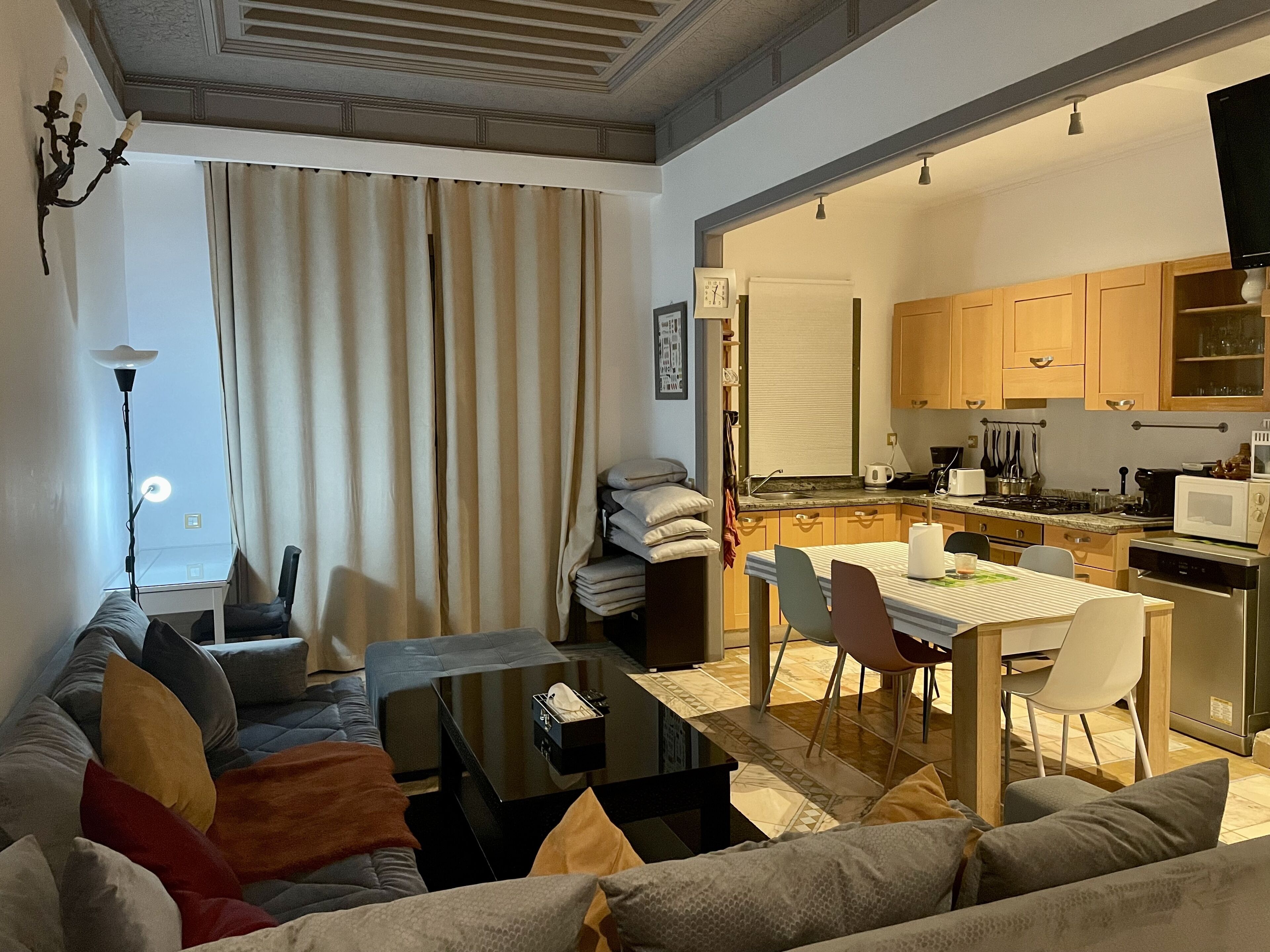 A living room with a sofa, a coffee table, and a dining area with chairs and a table. There is a kitchen visible through an open doorway.