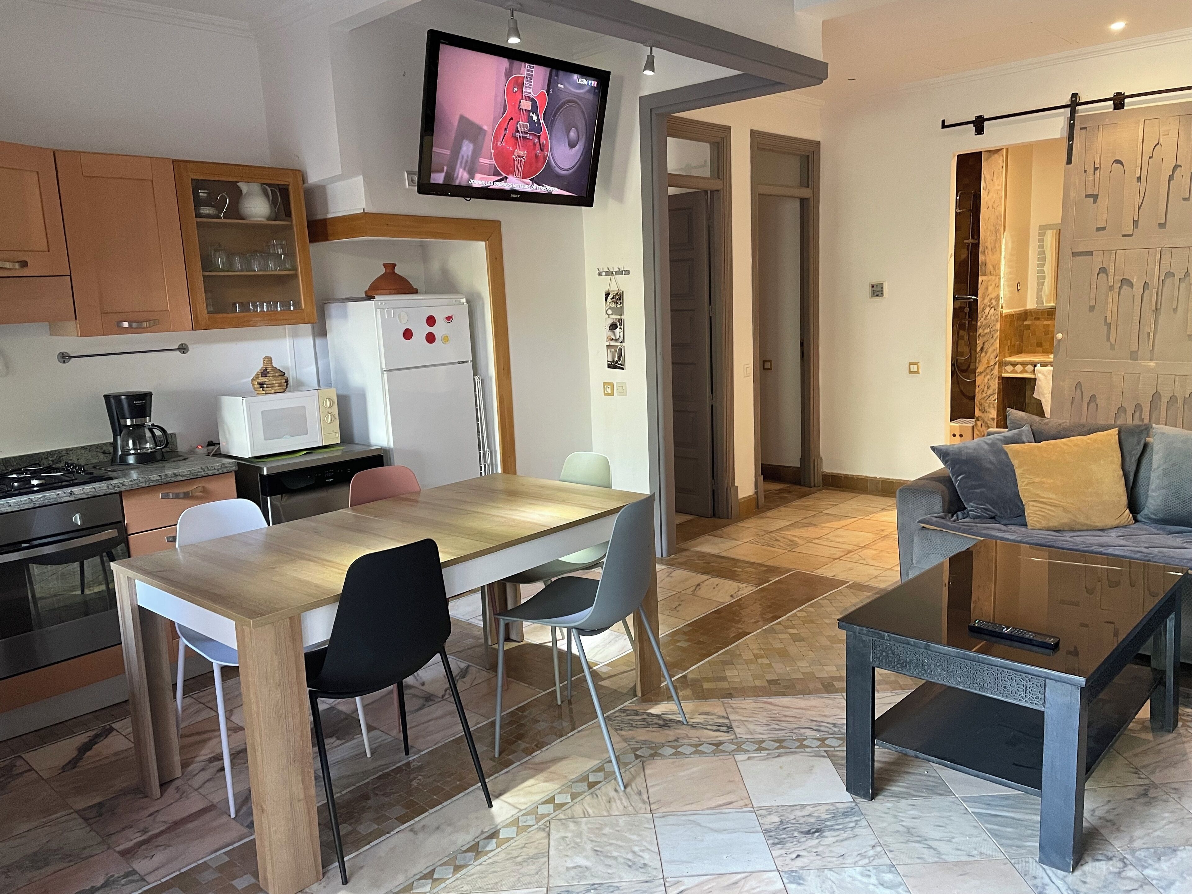 A modern kitchen with a dining area, a TV mounted on the wall, and a living area with a sofa and a coffee table.