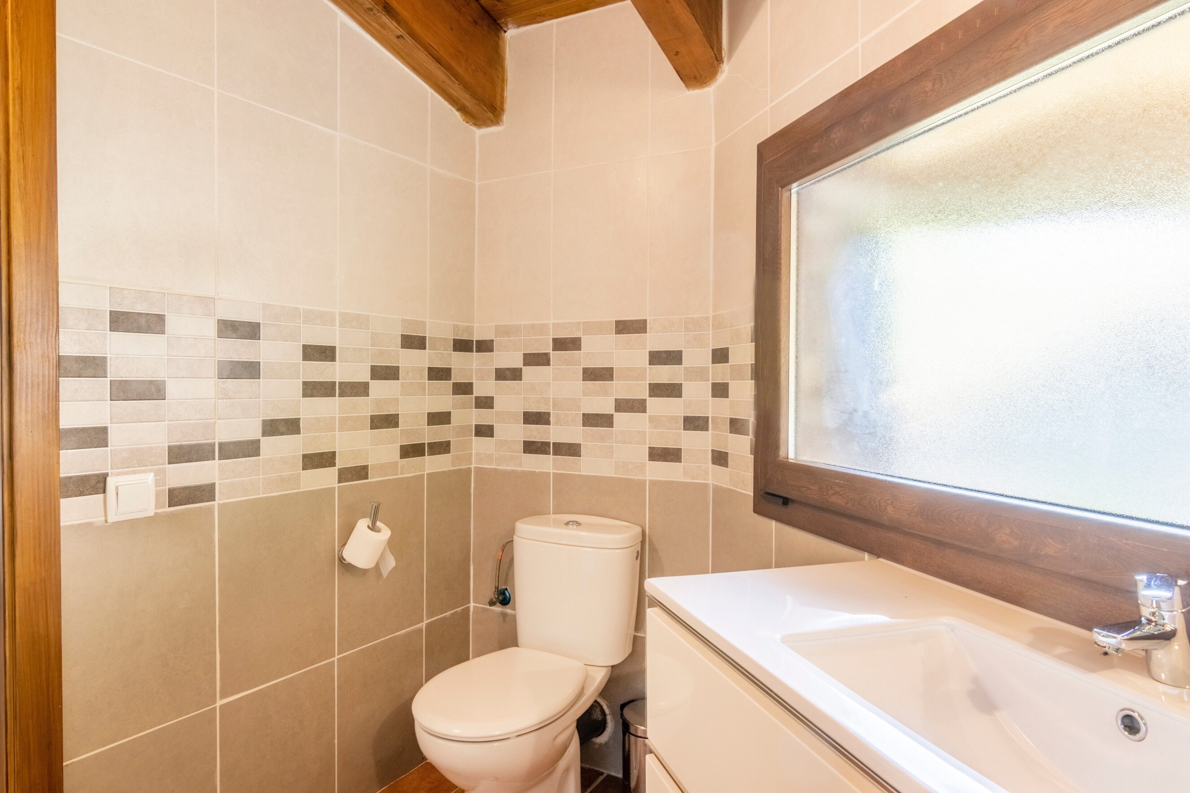 Un baño con pared de azulejos, inodoro, lavamanos y ventana.